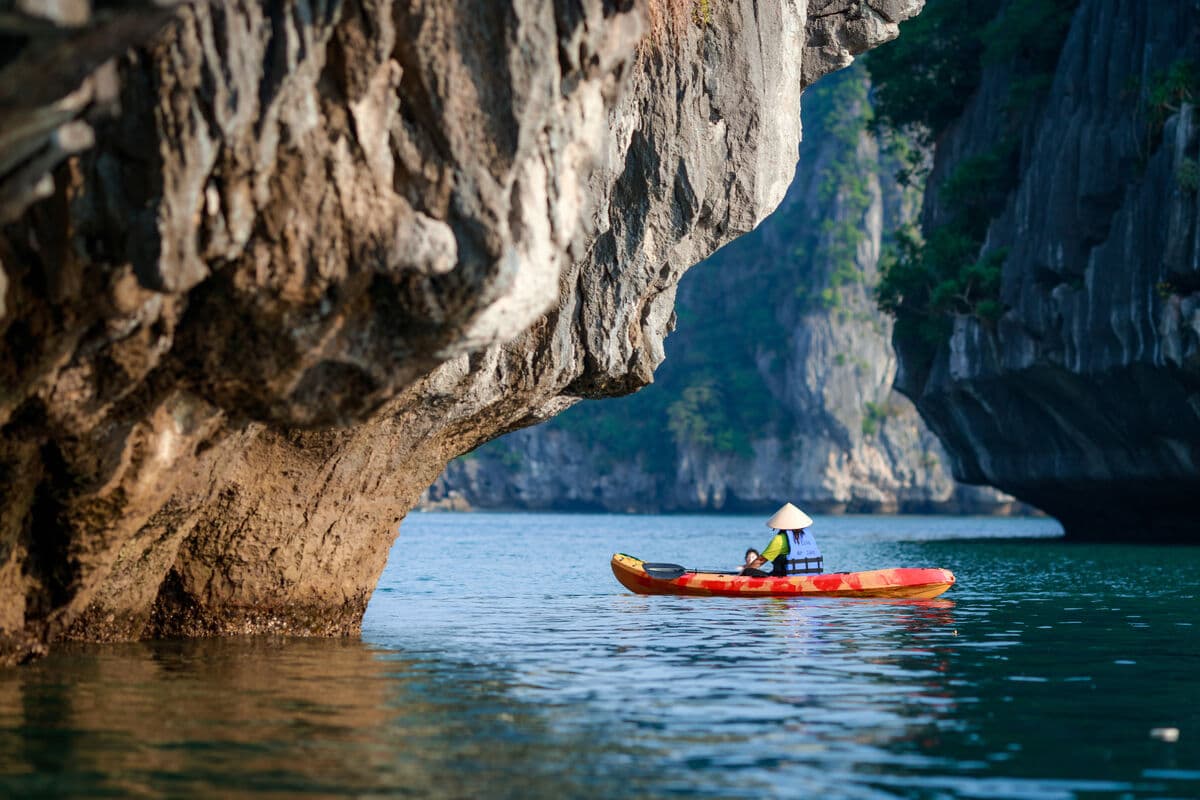Solo kayaker in a conical hat paddling beneath a limestone overhang